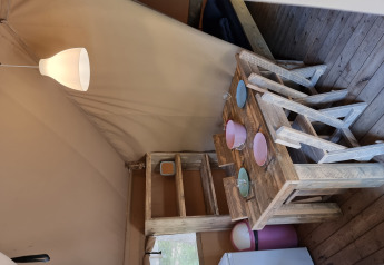 Intérieur d'une tente safari avec mobilier en bois rustique, table, chaises, vaisselle et lampe allumée.