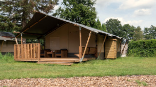 Foto van een safaritent met houten veranda, zitgedeelte en schuurtje in een groene, natuurlijke omgeving.