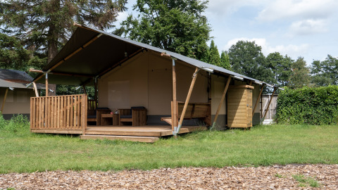 Photo d'une tente safari avec terrasse en bois, coin salon et cabanon dans un cadre naturel verdoyant.