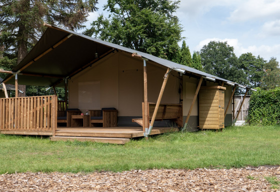 Photo d'une tente safari avec terrasse en bois, coin salon et cabanon dans un cadre naturel verdoyant.