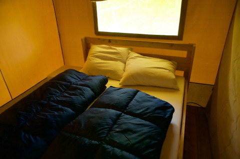 Vista interior de una tienda safari con una cama doble, almohadas y sacos de dormir bajo una gran ventana.