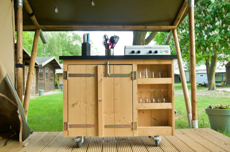 Outdoor wooden kitchen with gas stove, utensils, and glasses set under a safari tent structure.