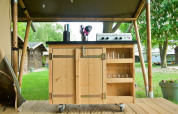 Cocina de madera al aire libre con estufa de gas, utensilios y vasos bajo una carpa tipo safari.