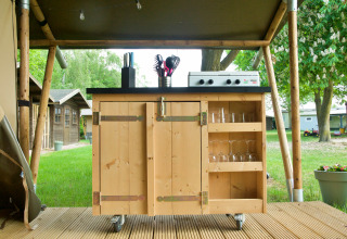Cocina de madera al aire libre con estufa de gas, utensilios y vasos bajo una carpa tipo safari.