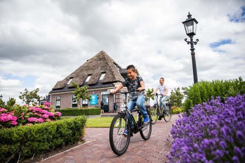 Due persone pedalano su un sentiero fiorito a Park Westerkogge con una tipica casa olandese sullo sfondo.