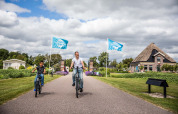 Far og søn cykler gennem ferieparken Park Westerkogge i Nordholland, Holland, under blå TopParken-flag.