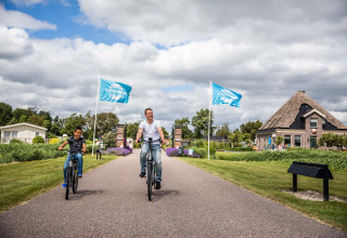 Vater und Sohn fahren Fahrrad im Ferienpark Park Westerkogge, Nordholland, Niederlande, bei TopParken-Flaggen.