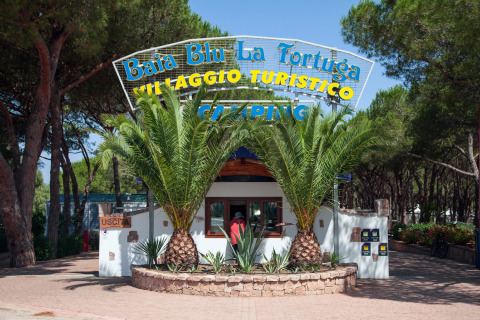 Entrada del Camping Village Baia Blu La Tortuga en Cerdeña, Italia, rodeada de palmeras y bosque de pinos.