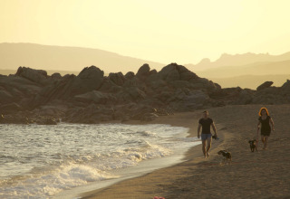 To personer og to hunde går en tur langs stranden ved solnedgang, Camping Village Baia Blu La Tortuga, Sardinien.