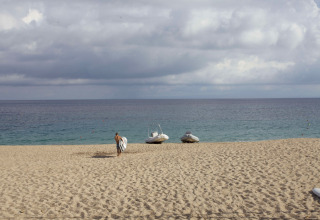 En strand med en mand, der bærer et surfbræt, to både på sandet og havet ved Camping Village Baia Blu La Tortuga.