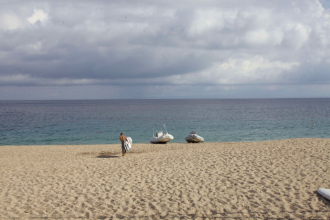 Een strand met een man en surfplank, twee boten op het zand bij Camping Village Baia Blu La Tortuga, Sardinië.