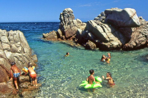 Niños juegan y nadan en agua clara entre rocas en Camping Village Baia Blu La Tortuga, Cerdeña, Italia.