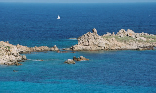 Mar azul claro con un velero a lo lejos cerca de una costa rocosa en los alrededores de Aglientu, Cerdeña, Italia.