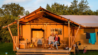 Pareja sentada afuera de una tienda safari con baño privado en Holiday Park Mölke, Países Bajos, al atardecer.
