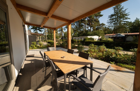 Overdækket terrasse ved en lodge med spisebord, seks stole og udsigt til hytter og grønne omgivelser.