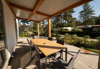 Overdekt terras bij een lodge met houten eettafel, zes stoelen en uitzicht op andere huisjes en groen.