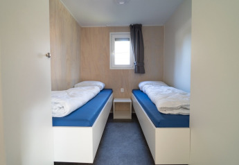 Chambre minimaliste d’un lodge avec deux lits simples, matelas bleus, couettes blanches et petite fenêtre.