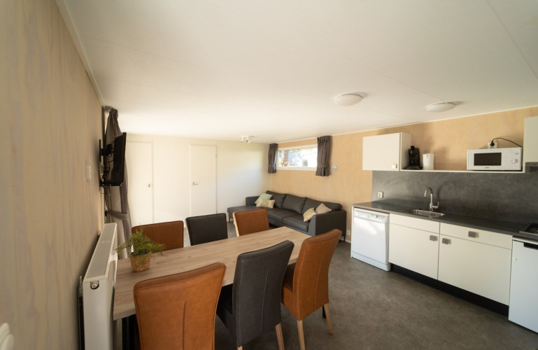 Interior luminoso y moderno de una cabaña con mesa de comedor, cocina equipada y cómodo sofá gris.