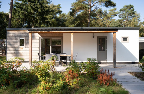 Lodge moderno con fachada de madera, terraza con mesa y sillas, rodeado de plantas y árboles altos.