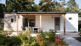 Lodge moderno con fachada de madera, terraza con mesa y sillas, rodeado de plantas y árboles altos.
