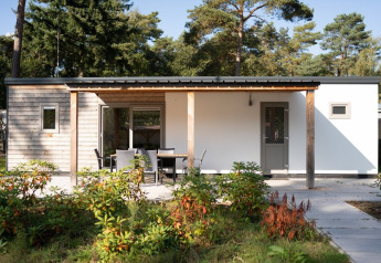 Lodge moderno con fachada de madera, terraza con mesa y sillas, rodeado de plantas y árboles altos.