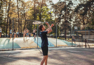 Junge spielt Tennis auf dem Platz bei Eurocamping Vessem BV, während Kinder hinten auf Hüpfkissen spielen.