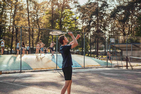 Ragazzo gioca a tennis a Eurocamping Vessem BV, con bambini che si divertono sul cuscino elastico dietro.