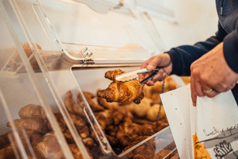 Personne prenant un croissant avec une pince dans une vitrine de boulangerie en libre-service du camping.