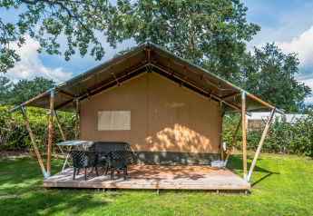 Safari-Zelt mit Holzterrasse, Gartentisch und Stühlen auf einer grünen Wiese, umgeben von Bäumen.