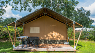 Safari tent met houten veranda, buitenstoelen en tafel, omgeven door gras en groene bomen.