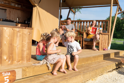 Familia disfrutando frente a SunLodge Taiga en Camping Cisano/San Vito, Italia, niños juegan en la terraza.