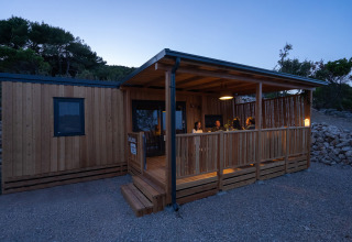 Vista nocturna de la SunLodge BigLeaf en Camping Cisano/San Vito, Italia, con personas en la terraza de madera.