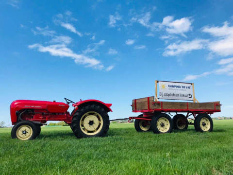 Tractor rojo con remolque y letrero de Camping 'de Val' sobre césped verde y cielo azul en Zeeland, Países Bajos.