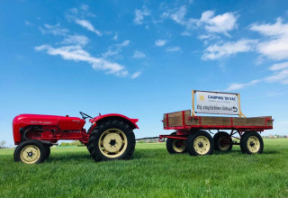 Tracteur rouge avec remorque et panneau Camping 'de Val' sur l’herbe verte sous ciel bleu en Zélande, Pays-Bas.