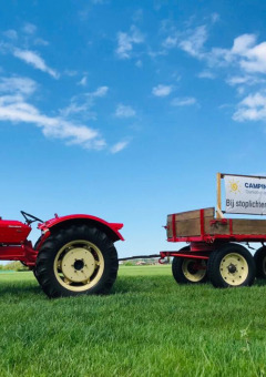 Red tractor with a trailer and sign for Camping 'de Val' on green grass and blue sky in Zeeland, Netherlands.