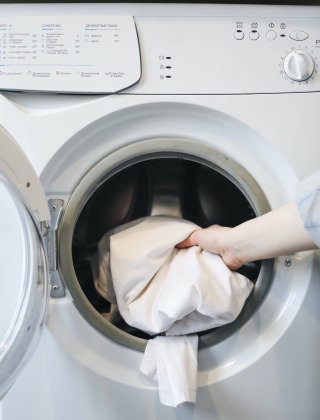 Eine Hand legt ein weißes Kleidungsstück in eine Frontlader-Waschmaschine in einer Glamping-Unterkunft.