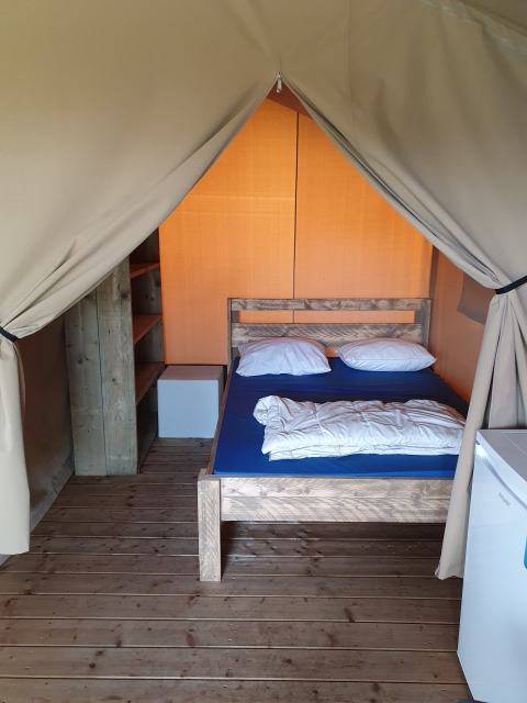 Interno di una tenda safari con pavimento in legno, letto matrimoniale, scaffali e piccolo frigo.