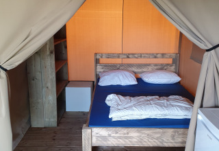 Interno di una tenda safari con pavimento in legno, letto matrimoniale, scaffali e piccolo frigo.
