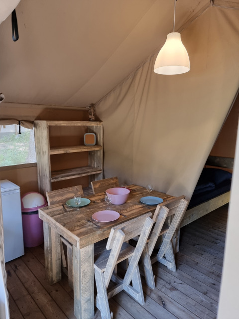 Interior de una tienda safari con mesa rústica de madera, cuatro sillas, estantería y luz acogedora.