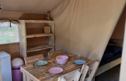 Interior de una tienda safari con mesa rústica de madera, cuatro sillas, estantería y luz acogedora.