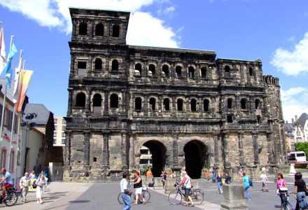 Personas caminando y en bicicleta junto a una antigua puerta romana de piedra en una plaza histórica y soleada.