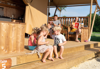Kinderen spelen op het terras van een safaritent, terwijl volwassenen achteraan een boek lezen.