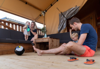 Familia relajándose en una tienda safari, con niños, balón de fútbol y sandalias en terraza de madera.