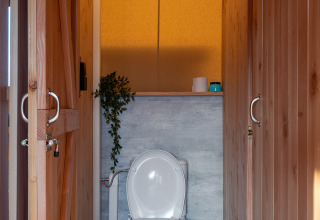 Toilettes modernes dans une tente safari, avec panneaux en bois, plante verte et poubelle métallique.