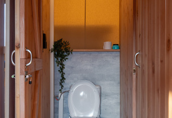 Modern toilet in een safaritent met houten panelen, groen plantje en metalen vuilbak aan de zijkant.