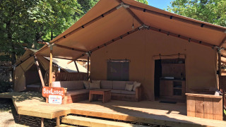 Safari tent called SunLodge Taiga&Kambi with a wooden deck, outdoor sofa, and green trees in the background.