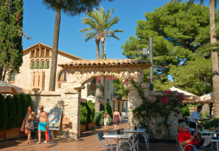 Scena all'aperto al Camping Vilanova Park in Catalogna, Spagna, con persone, tavoli e palme.