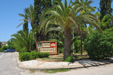Entrance sign for Camping Vilanova Park in Catalonia, Spain, surrounded by palm trees and lush greenery.