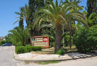 Entrance sign for Camping Vilanova Park in Catalonia, Spain, surrounded by palm trees and lush greenery.
