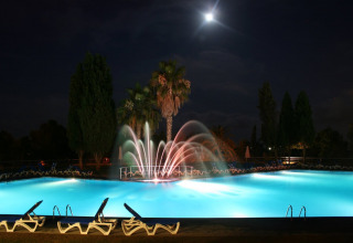 Oplyst swimmingpool med springvand og palmetræer om natten i Camping Vilanova Park, Catalonien, Spanien.